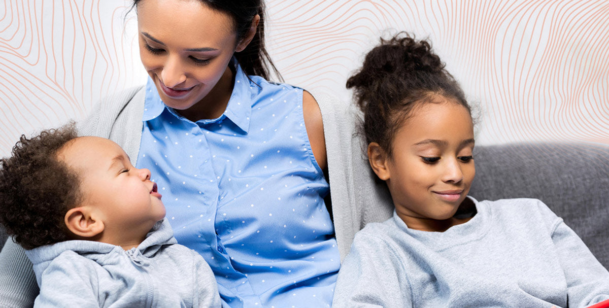A woman holding a baby, with a young girl sitting on a couch nearby.
