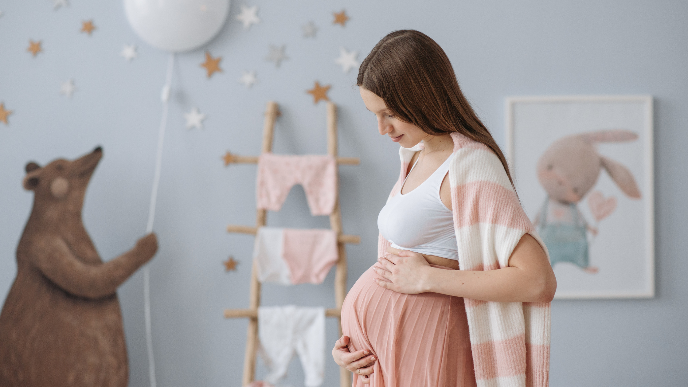 A pregnant woman standing indoors, holding her belly, with baby clothes hanging on a stick in the background.