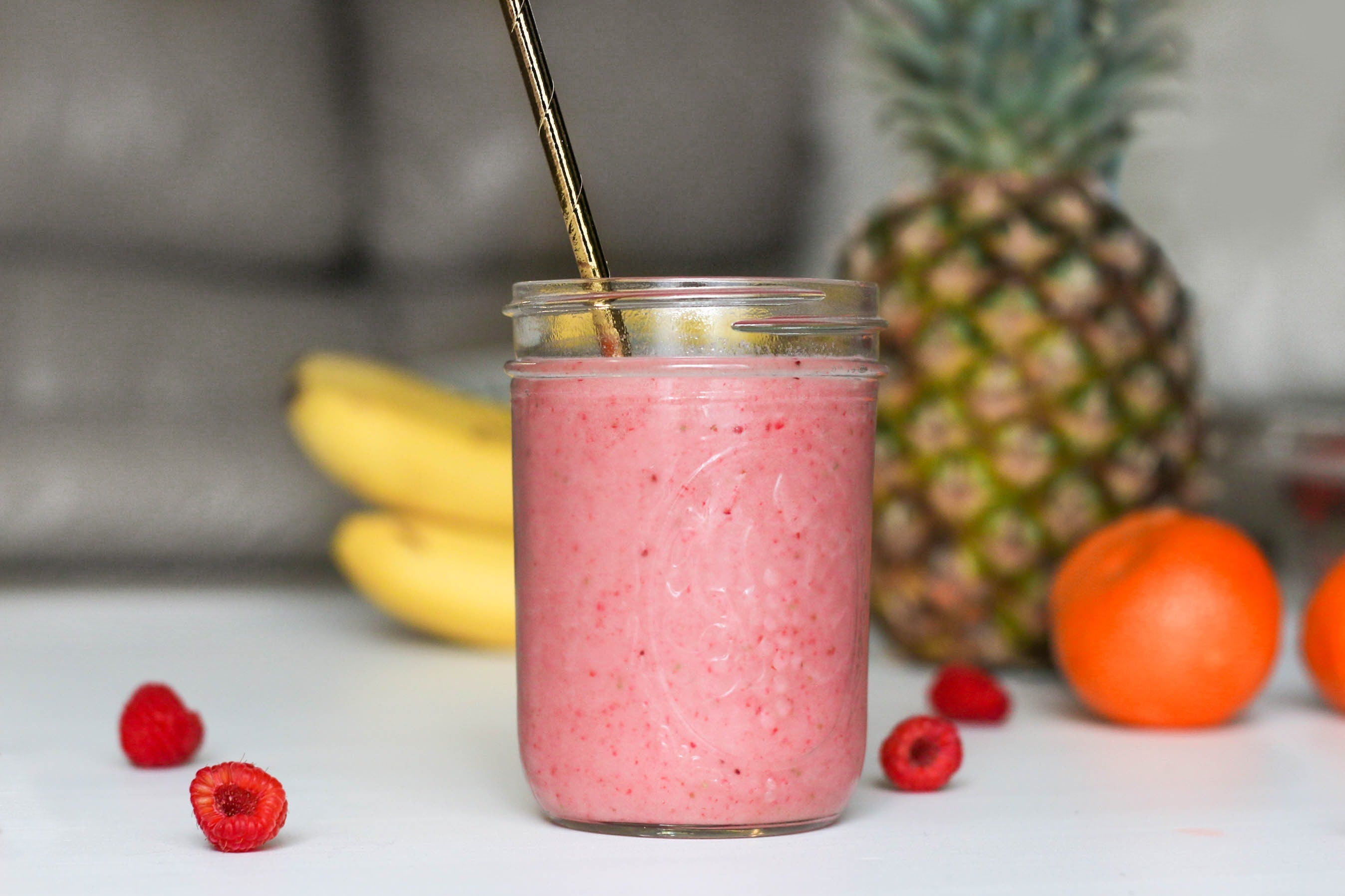 A pink smoothie in a jar with a gold spoon, surrounded by close-ups of an orange, bananas, raspberries, and a red berry.