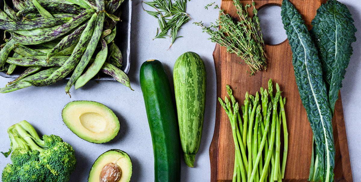 A variety of vegetables on a wooden surface, including broccoli, asparagus, beans, avocado halves, and leafy greens.
