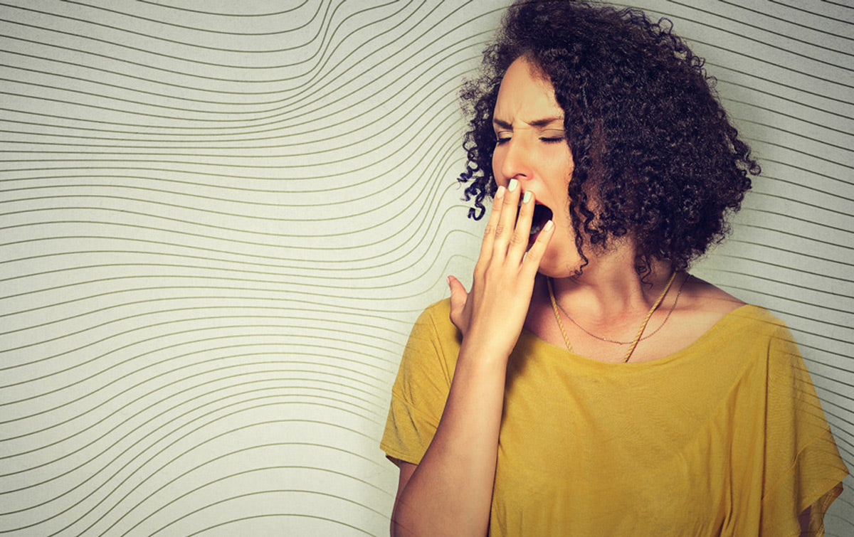 A woman in a yellow shirt yawning with her hand covering her mouth.
