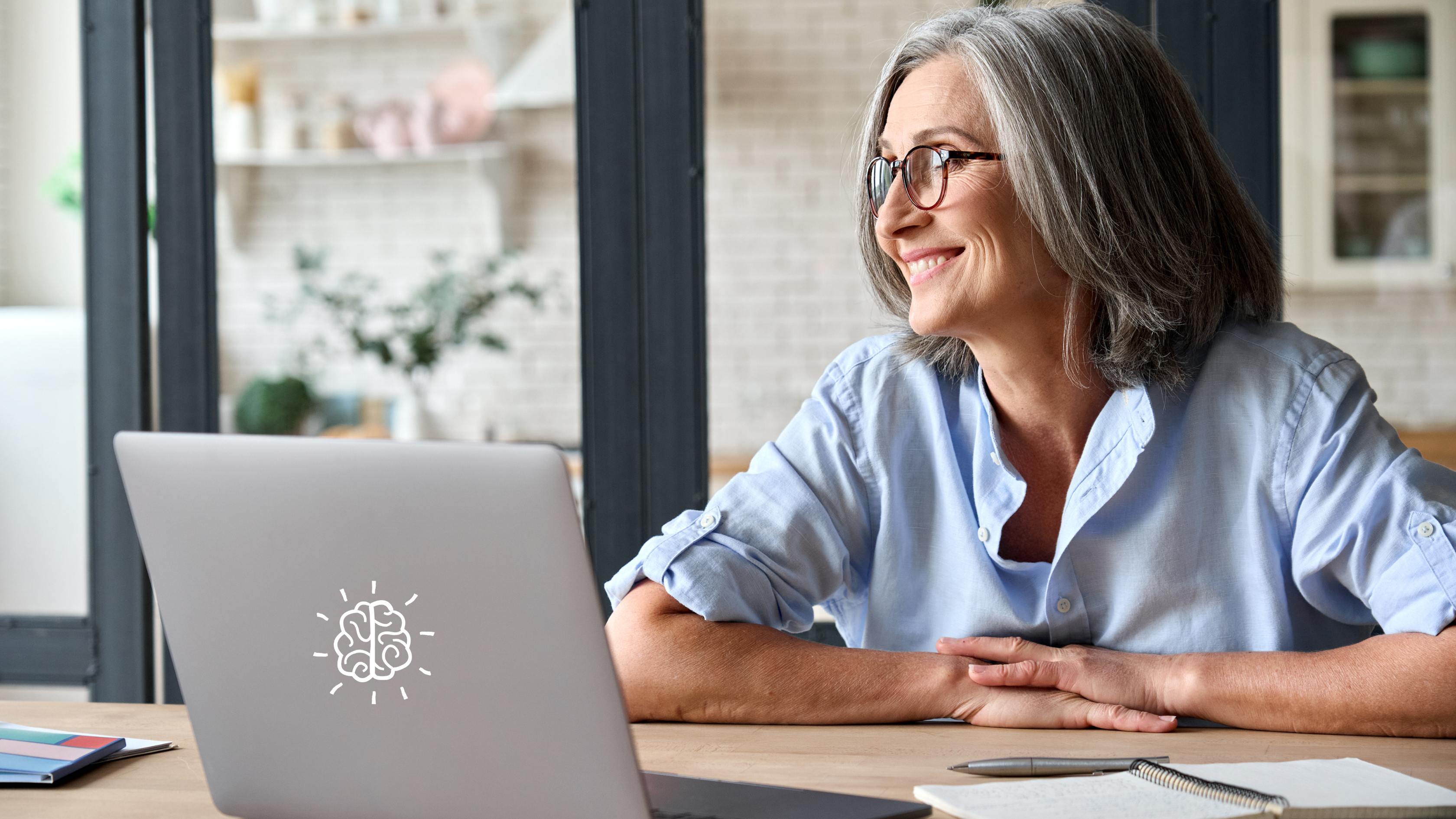 A smiling woman with grey hair and glasses sits at a desk, looking at a laptop with a brain design on its back.