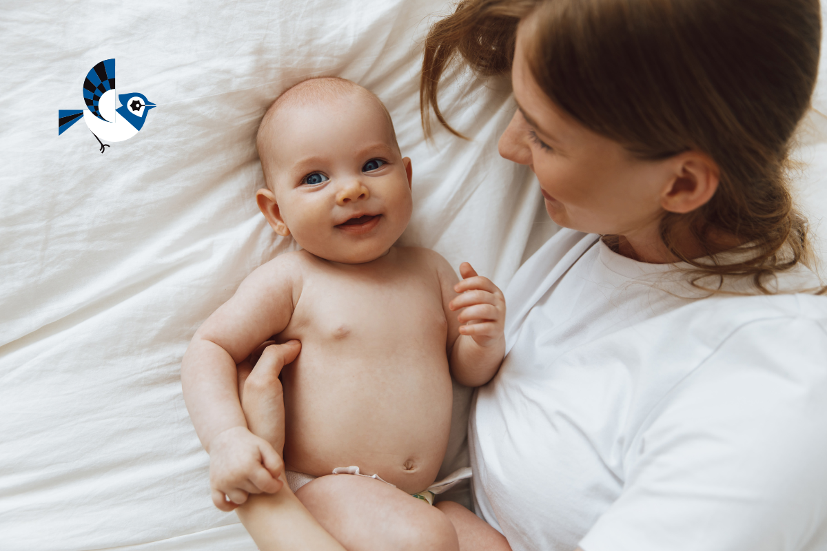 Smiling baby lying on a soft white bed, looking up at their mother, who looks down at the baby very lovingly. A warm and intimate moment between mom and baby.