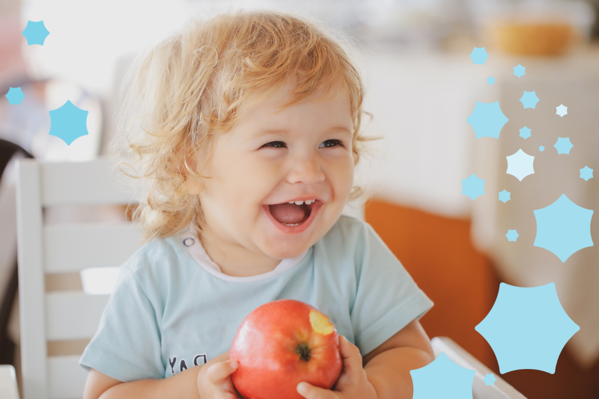 A happy toddler with curly blonde hair and a big smile, sitting at a table and holding a red apple. The child is wearing a light blue shirt, and the background is softly blurred. Blue star decorations are on the right side of the image.