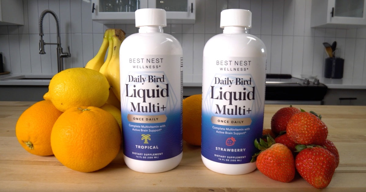 Bottles of Liquid Multi Plus on a counter, accompanied by close-ups of various fruits including oranges, strawberries, and bananas in the background.