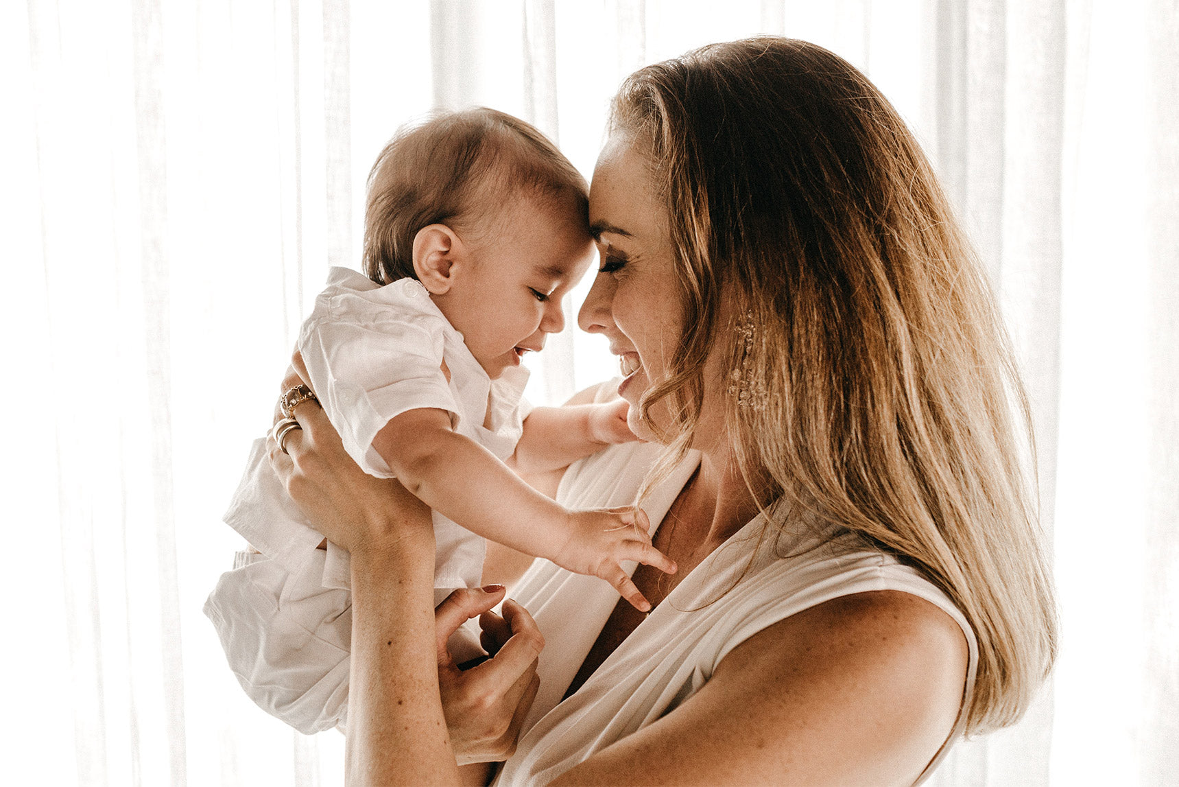 A woman smiling with her eyes closed, holding a baby close, depicting a loving, tender moment indoors.